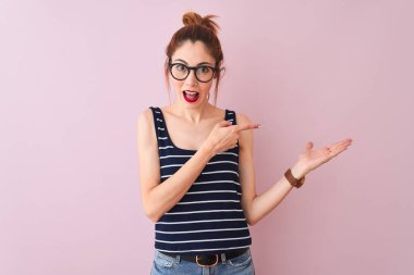 Kızıl saçlı, çizgili tişörtlü bir kadın izole edilmiş pembe arka planda dikiliyor kameraya hayret ediyor ve gülümsüyor..