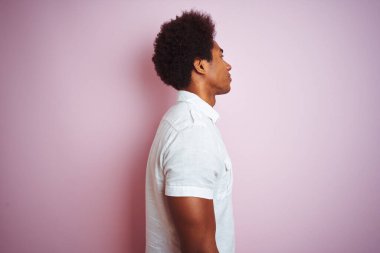 Afro saçlı, beyaz tişörtlü genç bir adam izole edilmiş pembe arka plan üzerinde duruyor. Yan yana bakıyor, rahat bir görünüm sergiliyor, kendine güvenen bir gülümsemeyle doğal bir yüz ifadesi sergiliyor..
