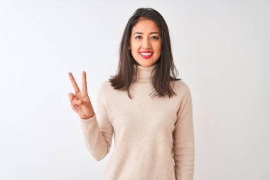 Balıkçı yaka kazak giyen güzel Çinli kadın izole edilmiş beyaz arka plan üzerinde duruyor ve parmaklarıyla iki numarayı gösterirken kendinden emin ve mutlu bir şekilde gülümsüyor..