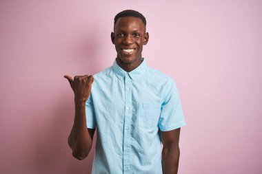 Mavi gömlekli Afro-Amerikan bir adam pembe arka planda dikilip gülümsüyor ve başparmağıyla yan tarafa bakıyor..
