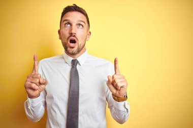 Sarı izole arka plan üzerinde zarif beyaz gömlek giyen genç yakışıklı iş adamı şaşırmış ve şaşırmış bir şekilde yukarı bakıyor ve parmaklarıyla işaret ediyor ve kollarını kaldırıyordu..