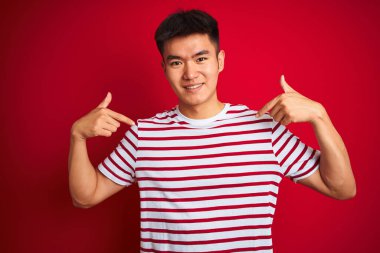 Asyalı genç bir Çinli, çizgili tişört giyiyor. İzole edilmiş kırmızı arka planda duruyor. Yüzünde bir gülümsemeyle, gururlu ve mutlu bir şekilde kendini gösteriyor..