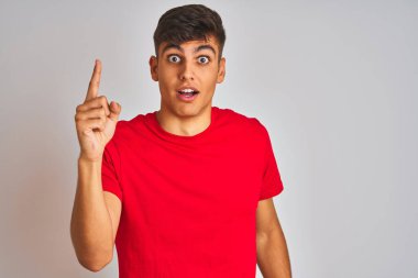 İzole edilmiş beyaz arka planda duran kırmızı tişörtlü genç Hintli adam başarılı bir fikirle işaret ediyor. Heyecanlı ve mutlu. Bir numara..