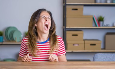 Orta yaş yaşlı kadın evde masada oturan kızgın ve deli çığlık sinirli ve öfkeli, öfke ile bağırarak. Öfke ve agresif kavram.