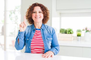 Orta yaşlı, kıvırcık saçlı, kot ceket giyen bir numaralı parmağını gösterip mutlu ve kendinden emin bir şekilde gülümseyen bir kadın..