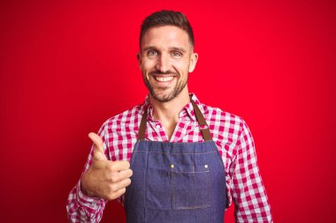 Kırmızı izole arka planda dükkan sahibi önlüğü giyen yakışıklı genç adam mutlu bir el hareketi yapıyor. Kameraya bakıp başarıyı gösteren ifade onaylanıyor.