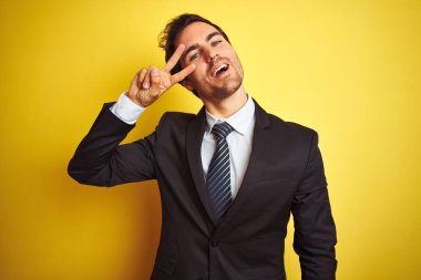 Takım elbise ve kravat giyen genç yakışıklı işadamı izole sarı arka plan üzerinde duran yüz üzerinde parmakları ile barış sembolü yapıyor, zafer gösteren neşeli gülümseyen