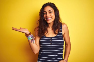 İzole sarı arka plan üzerinde duran çizgili t-shirt giyen genç güzel kadın neşeli sunan gülümseyerek ve kameraya bakarak avuç içi ile işaret.