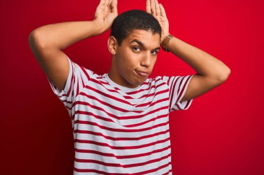 İzole edilmiş kırmızı arka plan üzerinde çizgili tişört giyen genç, yakışıklı Arap adam avuçlarında alaycı ve şüpheci bakışlarla tavşan kulağı hareketi yapıyor. Paskalya tavşanı kavramı.