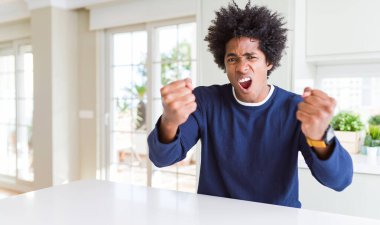 Genç Afro-Amerikalı bir adam rahat bir süveter giyip kızgın ve kızgın bir şekilde evde oturmuş öfkeyle bağırırken yumruklarını kaldırıyordu. Öfke ve agresif kavram.