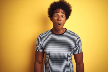 Afro saçlı, donanma çizgili tişörtlü, izole edilmiş sarı arka planı olan afro-amerikalı bir adam korku, korku ve heyecan dolu bir yüz ifadesinden korkuyor ve şok oluyordu..