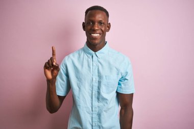 Mavi gömlekli Afro-Amerikalı bir adam pembe arka planda dikiliyor ve bir numaralı parmağını gösterirken kendinden emin ve mutlu bir şekilde gülümsüyor..