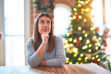 Genç ve güzel bir kadın Noel ağacı ve dekorasyonun etrafında masada oturmuş soru sormayı, düşünceli ifadeyi düşünüyor. Düşünceli bir yüzle gülümsüyor. Şüphe kavramı.