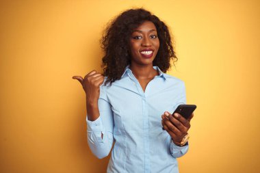 Genç Afro-Amerikan kadın akıllı telefon kullanıyor. İzole edilmiş sarı arka plan üzerinde duruyor ve gülümseyen bir gülümsemeyle baş parmağıyla onu işaret ediyor.