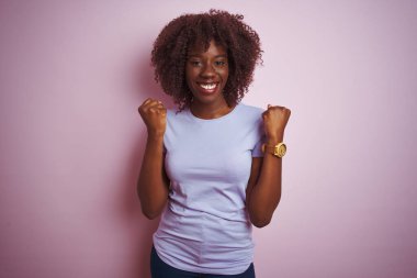 İzole edilmiş pembe arka planda duran tişört giyen Afrikalı genç bir kadın kollarını kaldırarak ve açık gözlerle başarıyı kutluyor ve şaşırıyor. Kazanan konsept.