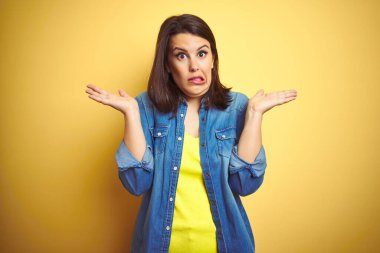 Sarı izole arka plan üzerine sıradan kot ceket giyen genç ve güzel bir esmer kadın. Kolları ve elleri havada, kafası karışmış bir yüz ifadesi. Şüphe kavramı.