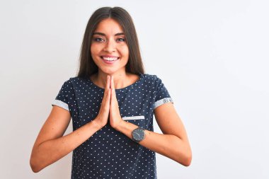 Genç, güzel bir kadın, mavi bir tişört giyiyor, izole edilmiş beyaz arka planda duruyor, ellerini birleştirip dua ediyor, af diliyor, kendinden emin bir şekilde gülümsüyor..