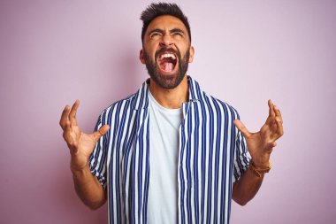 Genç, yakışıklı Hintli, yaz çizgili tişört giyen, izole edilmiş pembe arka planda deli deli deli bağırıp çağıran, agresif ifadeli ve kollarını kaldırmış genç bir adam. Hayal kırıklığı konsepti.