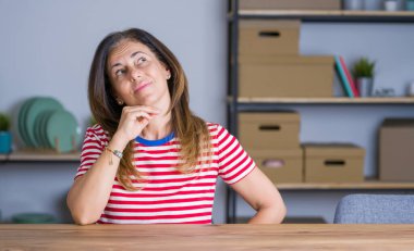 Orta yaş yaşlı kadın evde masada otururken elini çeneüzerinde sorgular, dalgın ifade. Düşünceli bir yüzle gülümsüyor. Şüphe kavramı.