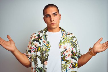Tatilde olan yakışıklı genç bir adam beyaz arka plan üzerine Hawaii gömleği giyiyor ve elleri ve kolları havada kafası karışmış bir ifade takınıyor. Şüphe kavramı.