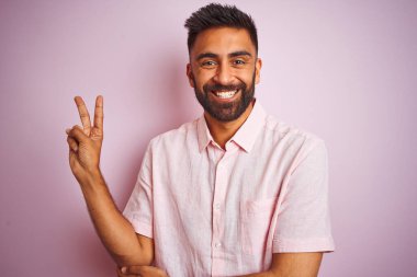 Izole pembe arka plan üzerinde duran rahat gömlek giyen genç hintli adam mutlu yüz ile gülümseyerek zafer işareti yapıyor kameraya göz kırparak. İki numara.