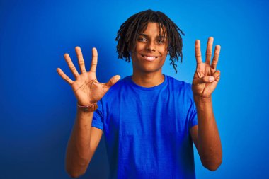Rastalı tişörtlü Afro-Amerikalı adam izole edilmiş mavi arka planda dikiliyor ve 8 numarayı işaret ediyor kendinden emin ve mutlu gülümserken..