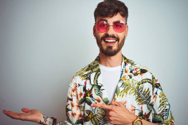 Tatilde dövmeli, güneş gözlüğü takan, izole edilmiş beyaz arka planda hayrete düşmüş ve kameraya gülümseyen bir adam..