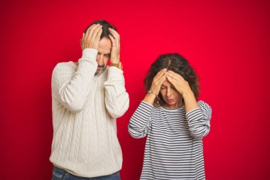Güzel orta yaşlı çift, izole edilmiş kırmızı arka plan üzerine kazak giyip acı ve migren yüzünden çaresiz ve stresli bir baş ağrısı çekiyor. Ellerini başının üstüne koy..