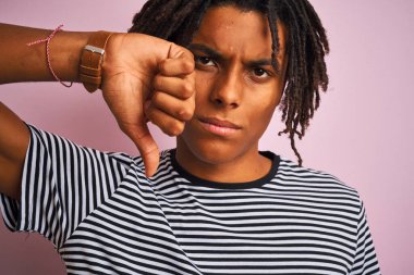 Rasta desenli tişörtlü Afro-Amerikalı adam izole edilmiş pembe arka planda kızgın yüzlü, olumsuz işaretli baş parmakları aşağıda, reddedilme konsepti var.
