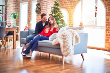 Young beautiful couple smiling happy and confident. Sitting on the sofa hugging around christmas tree at home