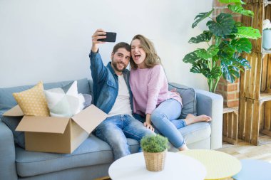 Yeni bir eve taşındığı için çok mutlu gülümseyen kanepede oturan akıllı telefon kullanarak selfie fotoğrafı çeken genç güzel çift
