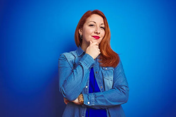 Young beautiful redhead woman wearing denim shirt standing over blue isolated background looking confident at the camera smiling with crossed arms and hand raised on chin. Thinking positive.