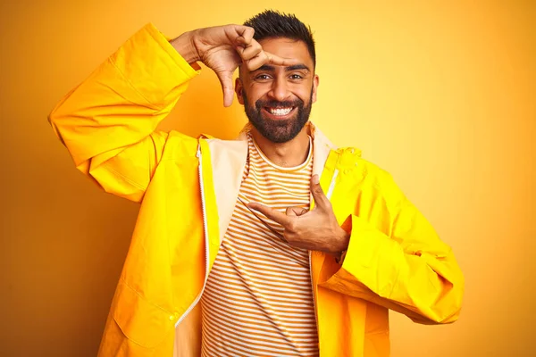Young indian man wearing raincoat standing over isolated yellow background smiling making frame with hands and fingers with happy face. Creativity and photography concept.