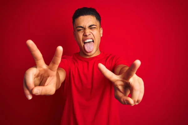 Tシャツを着た若いブラジル人男性が孤立した赤い背景の上に立っていて 両手の指が勝利サインをしているのを舌で笑っています 第二番 — ストック写真