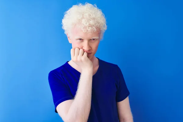Young albino blond man wearing casual t-shirt standing over isolated blue background looking stressed and nervous with hands on mouth biting nails. Anxiety problem.