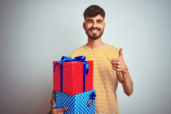 Young man with tattoo holding birthday gifts standing over isolated white background happy with big smile doing ok sign, thumb up with fingers, excellent sign