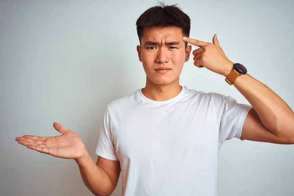 Young asian chinese man wearing t-shirt standing over isolated white background confused and annoyed with open palm showing copy space and pointing finger to forehead. Think about it.