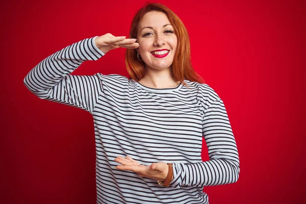 Young redhead woman wearing strapes navy shirt standing over red isolated background gesturing with hands showing big and large size sign, measure symbol. Smiling looking at the camera. Measuring concept.