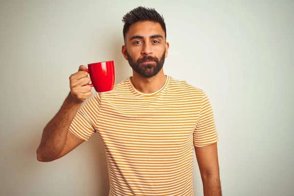 Young indian man drinking red cup of coffee standing over isolated white background with a confident expression on smart face thinking serious