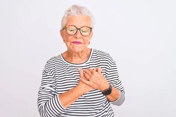 Senior grey-haired woman wearing striped navy t-shirt glasses over isolated white background smiling with hands on chest with closed eyes and grateful gesture on face. Health concept.