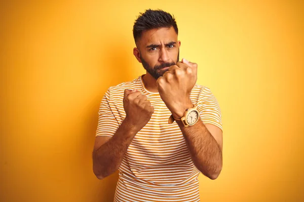 Young indian man wearing t-shirt standing over isolated yellow background Ready to fight with fist defense gesture, angry and upset face, afraid of problem