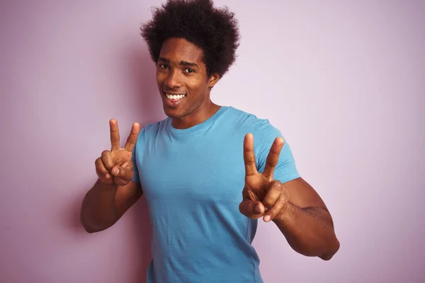 青いTシャツを着たアフロの髪をしたアフリカ系アメリカ人男性がピンクの背景の上に立って 勝利のサインをしている指を示すカメラを見て笑っている 第二番 — ストック写真