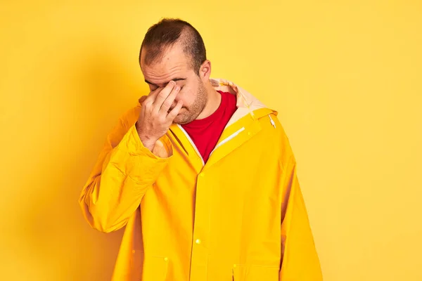 孤立した黄色の背景に立ってレインコートを着て若い男が疲れ鼻と目の疲労や頭痛を感じて摩擦疲れている ストレスとフラストレーションの概念 — ストック写真