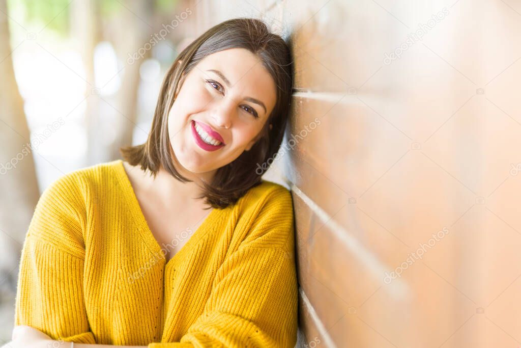 Beautiful young woman smiling confident and cheerful leaning on wooden wall, walking on the street of the city on a sunny day