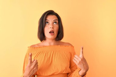 İzole edilmiş turuncu arka plan üzerinde duran gündelik tişört giyen genç ve güzel bir kadın şaşırmış ve şaşırmış bir şekilde yukarı bakıp parmaklarıyla işaret ediyor ve kollarını kaldırıyordu..