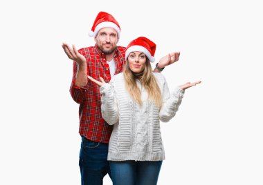 Genç çift aşık izole arka plan clueless ve şaşkın bir ifadeyle kollar ve eller kaldırdı üzerinde Noel şapka giyiyor. Şüphe kavramı.