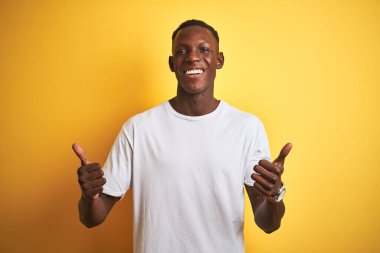Beyaz t-shirt giyen genç Afro-Amerikalı adam izole edilmiş sarı arka plan başarısı tabelasının üzerinde duruyor. Elleriyle olumlu jest yapıyor, gülümsüyor ve mutlu. Neşeli ifade ve kazanan jest.