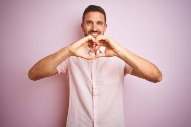 Pembe izole edilmiş arka planda zarif bir yaz gömleği giyen yakışıklı genç bir adam. Elleriyle kalp sembolü ve şekli gösteren bir aşkla gülümsüyor. Romantik konsept.