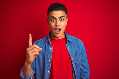 İzole edilmiş kırmızı arka planda duran kot gömlekli genç Brezilyalı adam başarılı bir fikri işaret ediyor. Heyecanlı ve mutlu. Bir numara..