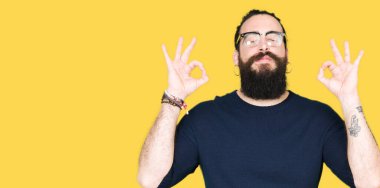 Uzun saç ve sakal gözlüklü genç hipster adamla rahatla ve gülümseyen gözlerle kapalı parmak jestle meditasyon yapıyor. Yoga kavramı.